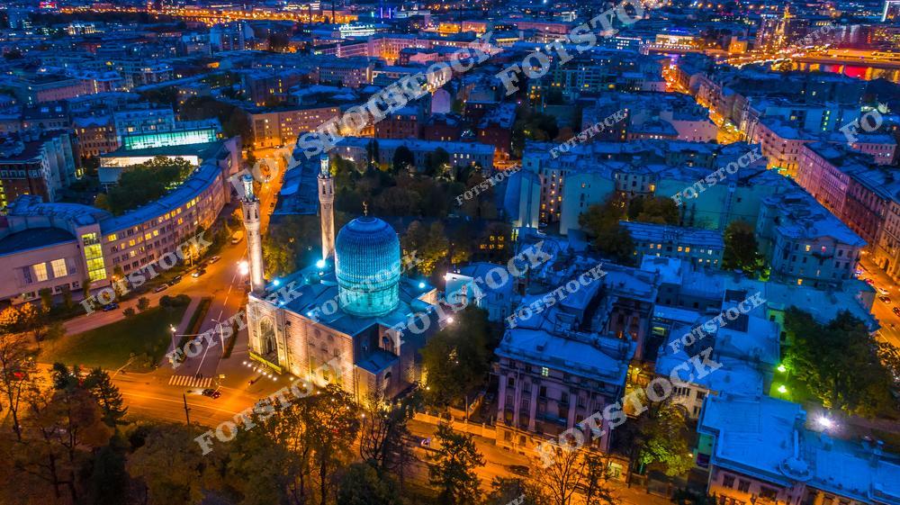 На фотографии изображена Санкт-Петербургская мечеть в вечернее время, окруженная огнями ночного города. Это одна из крупнейших мечетей Европы с ярко выраженными восточными архитектурными элементами, такими как высокие минареты и голубой купол. Подсветка придаёт мечети особое величие и подчеркивает её архитектурные детали. Вокруг мечети видны жилые кварталы и улицы, что делает вид уникальным сочетанием культур в центре Санкт-Петербурга.